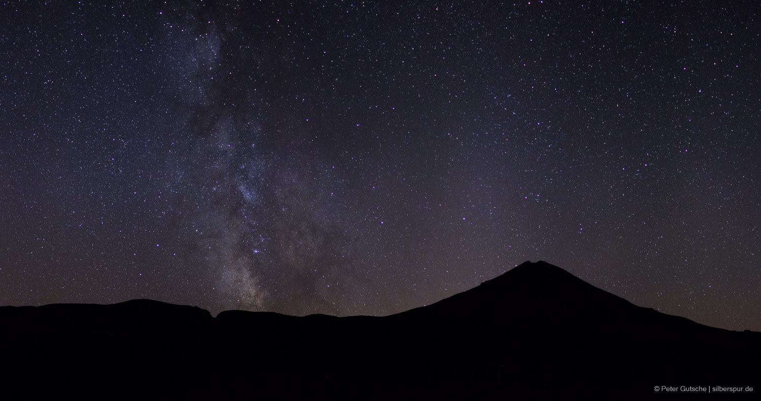 A dark mountain silhouette at night with the starry sky above.
The bright band of the Milky Way is clearly seen in the center of the image
as it rises perpendicular into the sky.