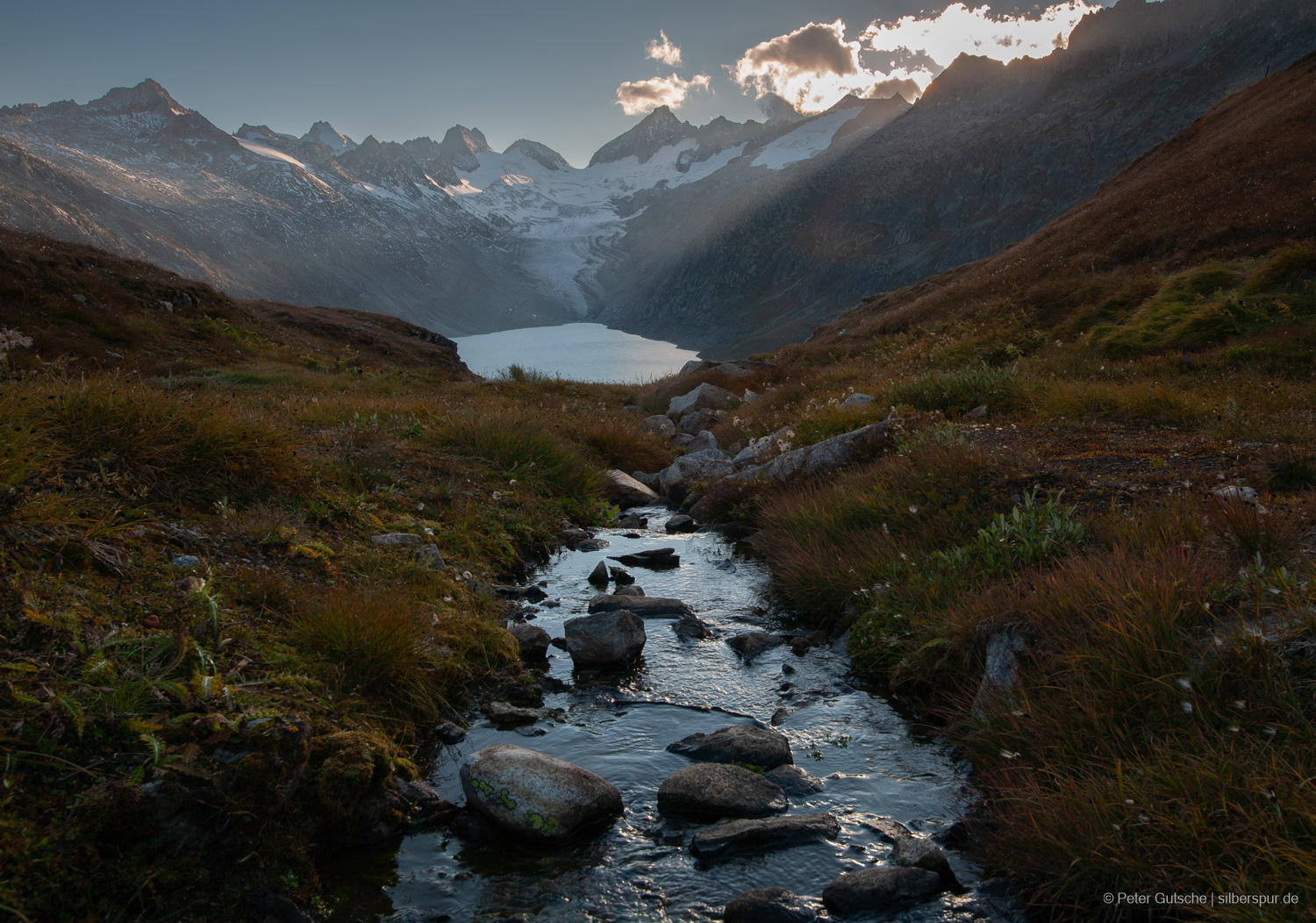 Ein glitzernder Gebirgsbach fließt vom Betrachter weg, direkt ins Gegenlicht der tief stehenden Nachmittagssonne. Die Wasseroberfläche reflektiert das Licht und lässt den Bach wie ein silbernes Band durch die alpine Landschaft ziehen. Im Hintergrund öffnet sich ein Tal mit einem See, der ruhig zwischen den Berghängen liegt. Dahinter ragt ein mächtiges Bergmassiv mit Gletscherfeldern auf; einzelne Gipfel zeichnen sich scharf gegen den hellen Himmel ab.