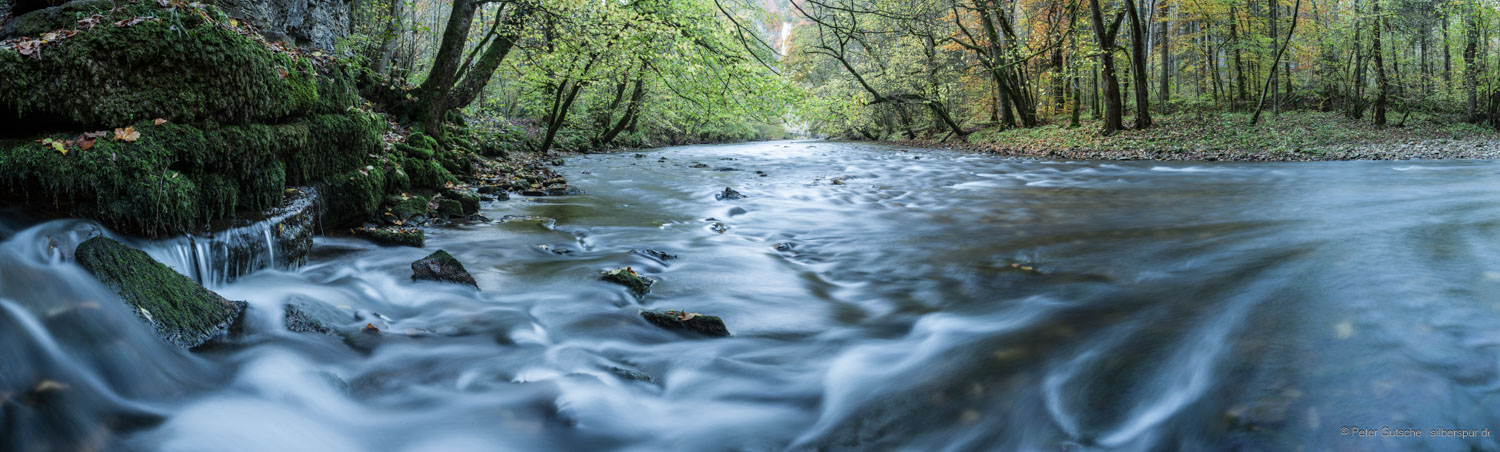 Panoramaaufnahme eines Flusses in herbstlicher Waldlandschaft. Im linken Vordergrund tritt Wasser aus Spalten im Kalkgestein aus und mündet in den Flusslauf. Die lange Belichtungszeit lässt die Wasseroberfläche weich und teils verschwommen erscheinen, was den Eindruck von Bewegung und Strömung verstärkt.