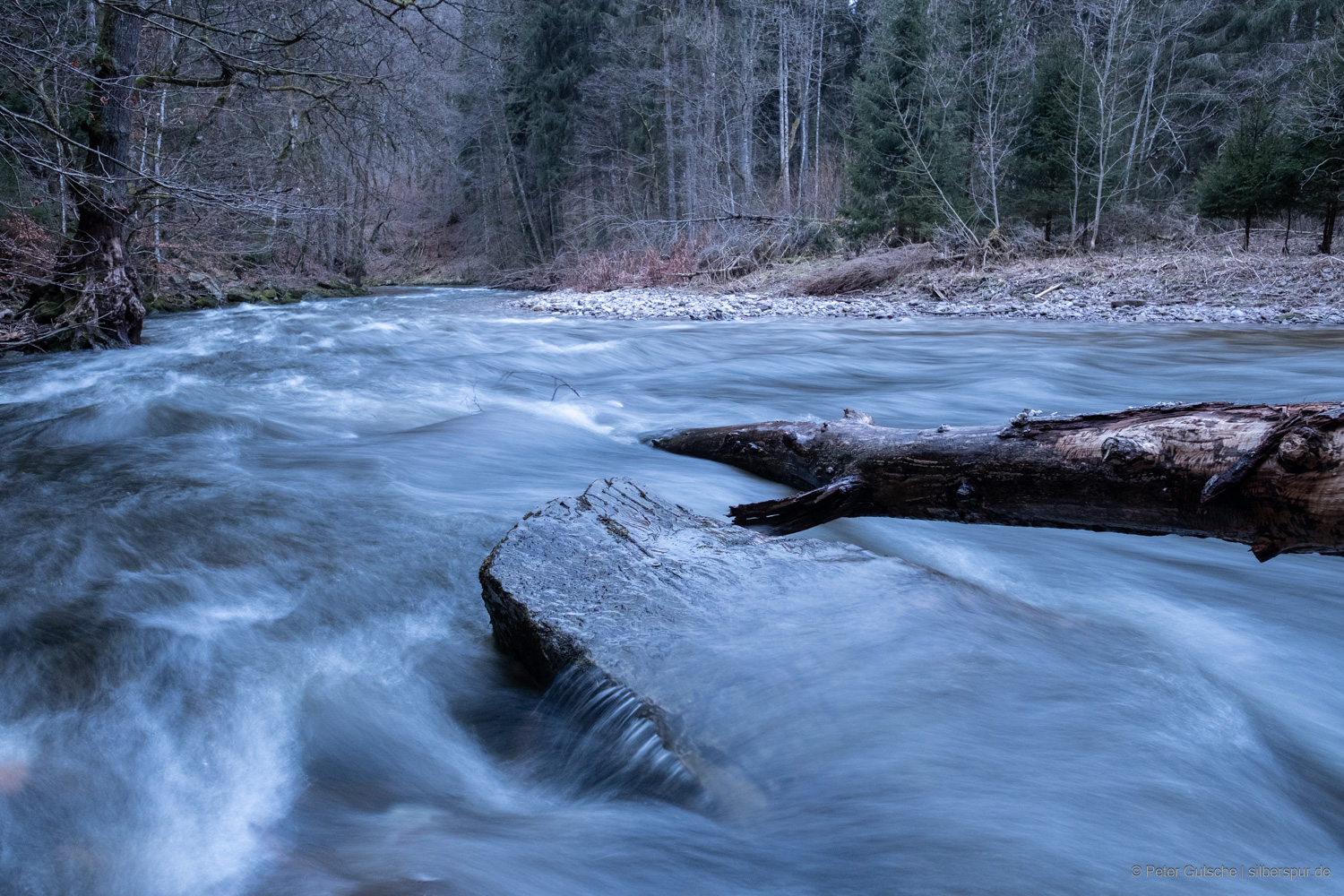 Ein strömender Fluss in Flussrichtung aufgenommen. Im Vordergrund taucht ein Fels teils aus der Strömung heraus und es ragt ein Baumstamm von hinten ins Bild. Durch die lange Belichtungszeit ist das Wasser verschwommen und wirkt fließend.