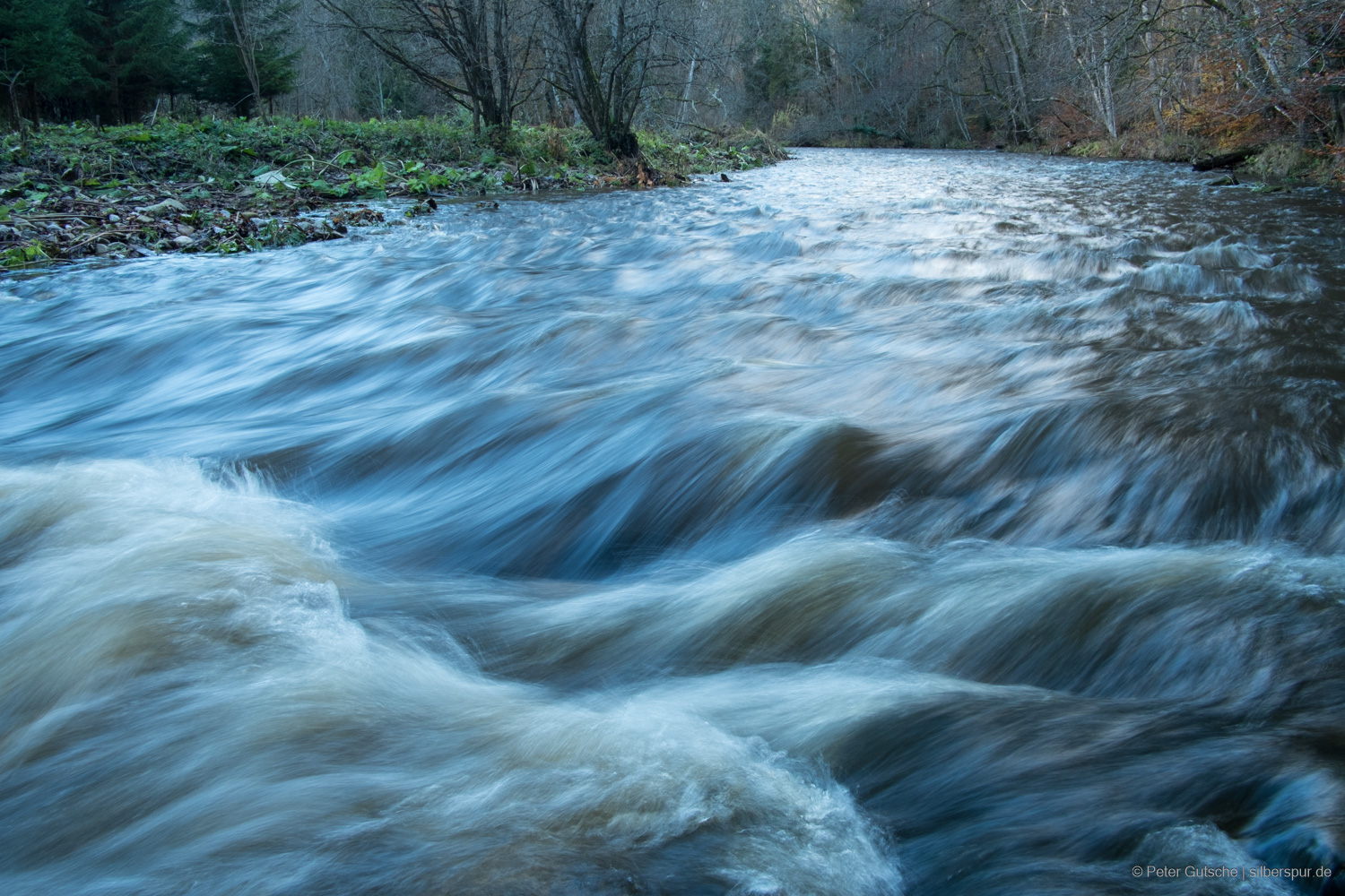 Der Fluss wurde aus niedriger Perspektive knapp oberhalb der Wasseroberfläche aufgenommen, und der der Blick führt flussaufwärts in die entgegengesetzte Strömungsrichtung. Durch die leicht verlängerte Belichtungszeit wirken die Wellen weichgezeichnet, das Wasser erscheint in strähnigen Strukturen, die die Dynamik der Strömung betonen.