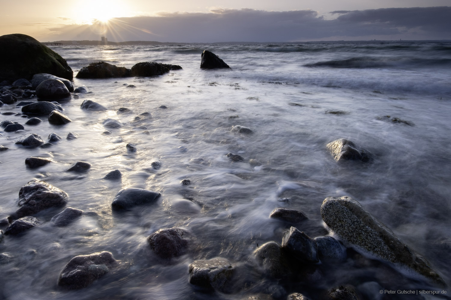 At the Baltic Sea beach during sunset, individual, sometimes large stones rise from the shallow water in the foreground. The incoming surf partially washes over them, making their wet surfaces shine brightly in the warm backlight. Due to the long exposure time, the water appears soft and blurred, giving the image a calm, almost meditative effect – despite the powerful movement of the waves.