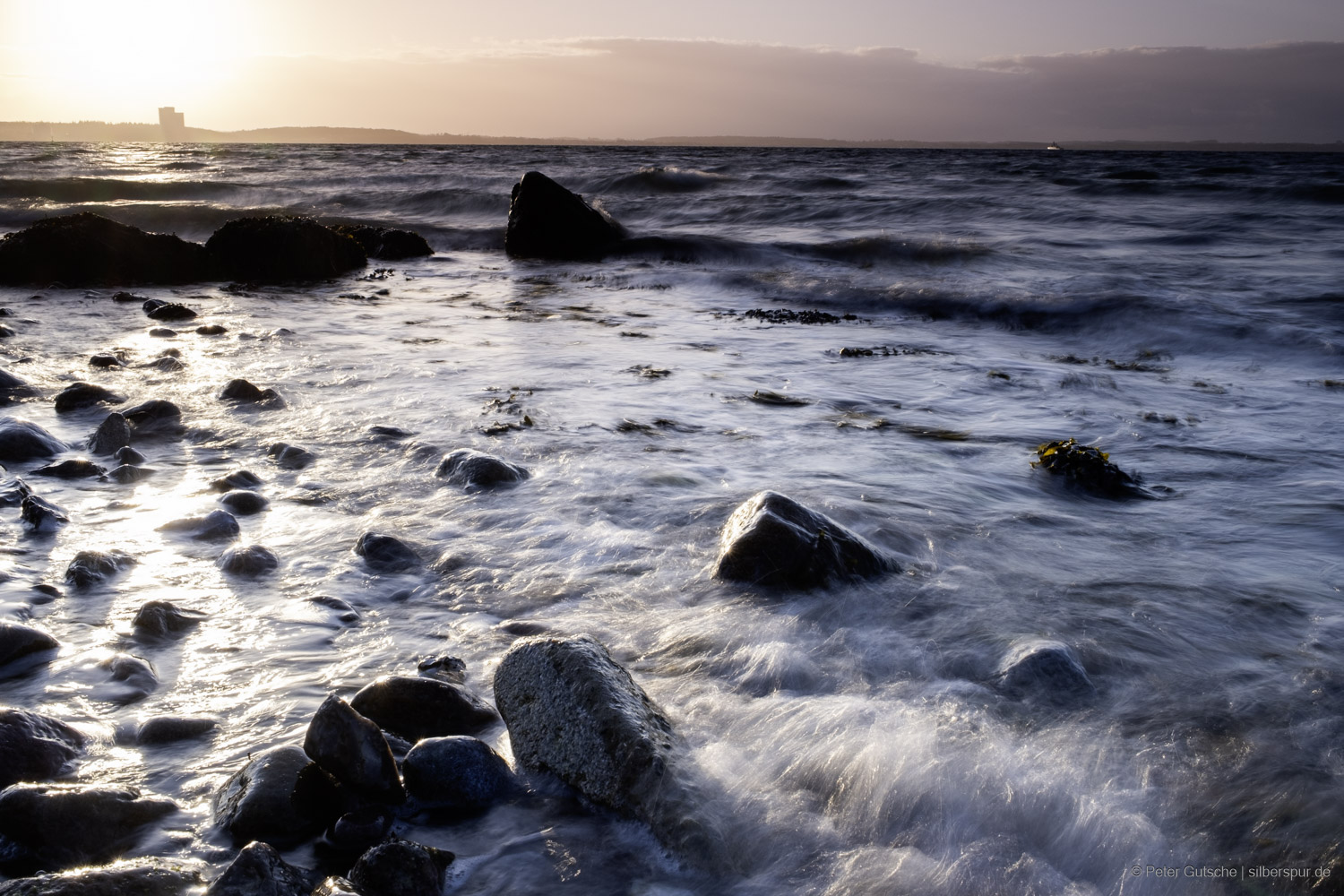 At the Baltic Sea beach during sunset, large stones jut out from the water in the foreground. The incoming waves wash over them, causing their wet surfaces to shine in the warm backlight. With a slightly shorter exposure time, the water remains softly blurred, yet individual water splashes and currents are now more clearly visible, sparkling like fine threads in the light of the setting sun. This creates a dynamic sense of movement while still maintaining the serene atmosphere of the scene.
