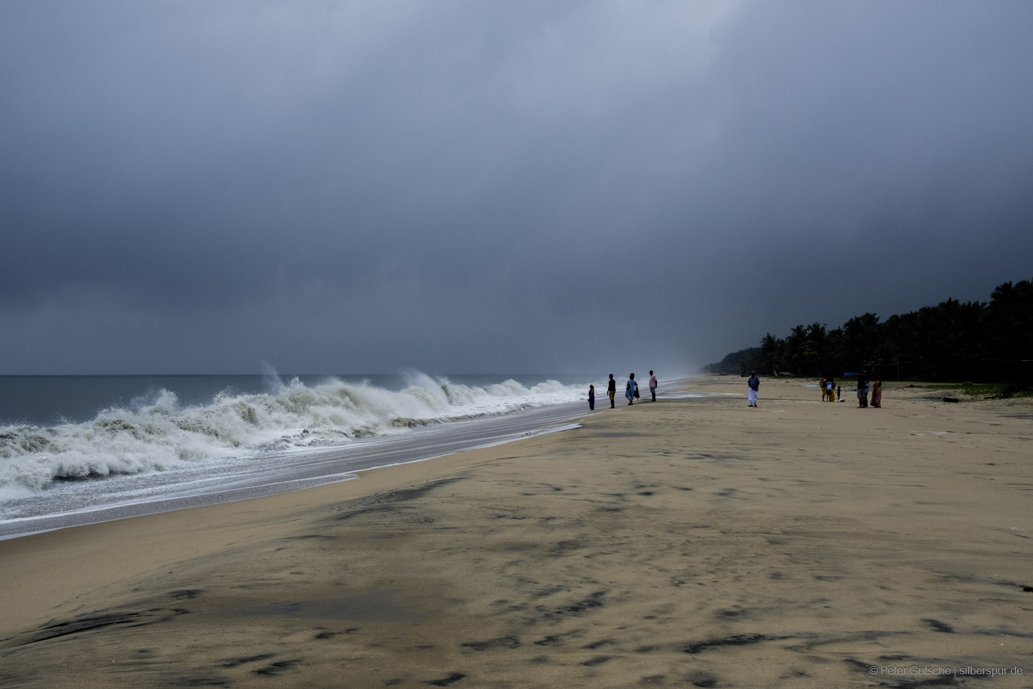 A dramatic beach scene on the coast of Kerala, South India. The sky is covered with dark clouds, adding a powerful and intense atmosphere to the scene. On the left, the roaring sea crashes with tall, white-capped waves onto the ochre-colored sandy beach, which stretches out toward the center. To the right, a dense palm forest is silhouetted in the twilight. In the distance, a group of people can be seen, some facing the sea. The waves rise so high in places that they tower over the people, highlighting the immense power of the ocean.