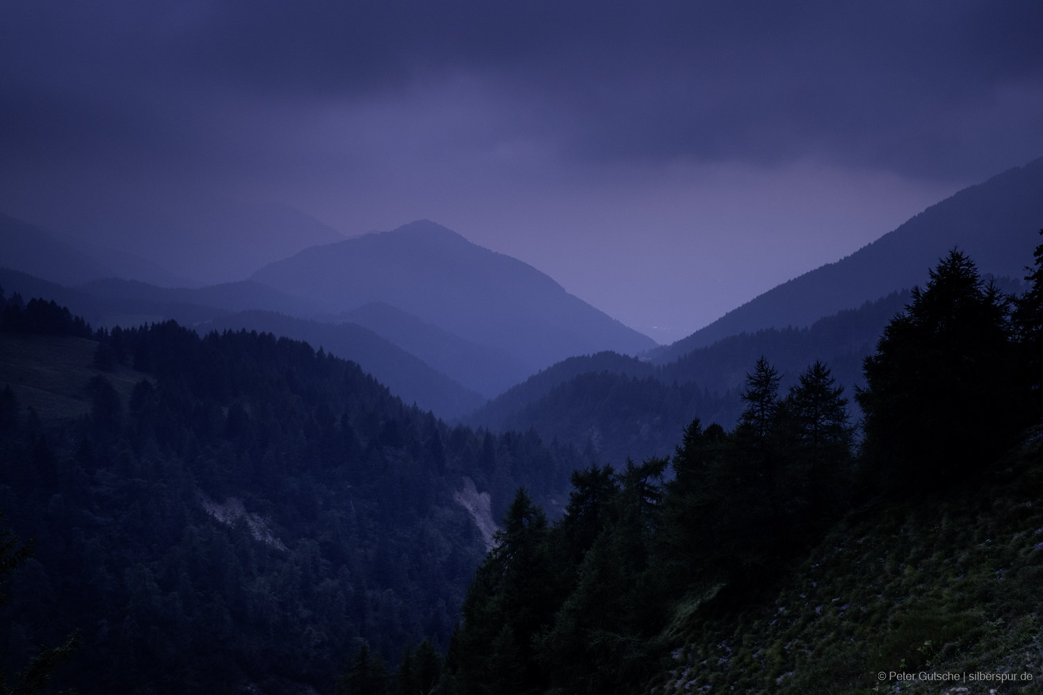 A mood-filled evening scene shows a deep blue mountain pass with a view into a vast valley. Several layered mountain ridges stretch into the distance, gradually fading in the misty evening light. Dark clouds hang above the scene, giving the landscape a mysterious and dramatic atmosphere.