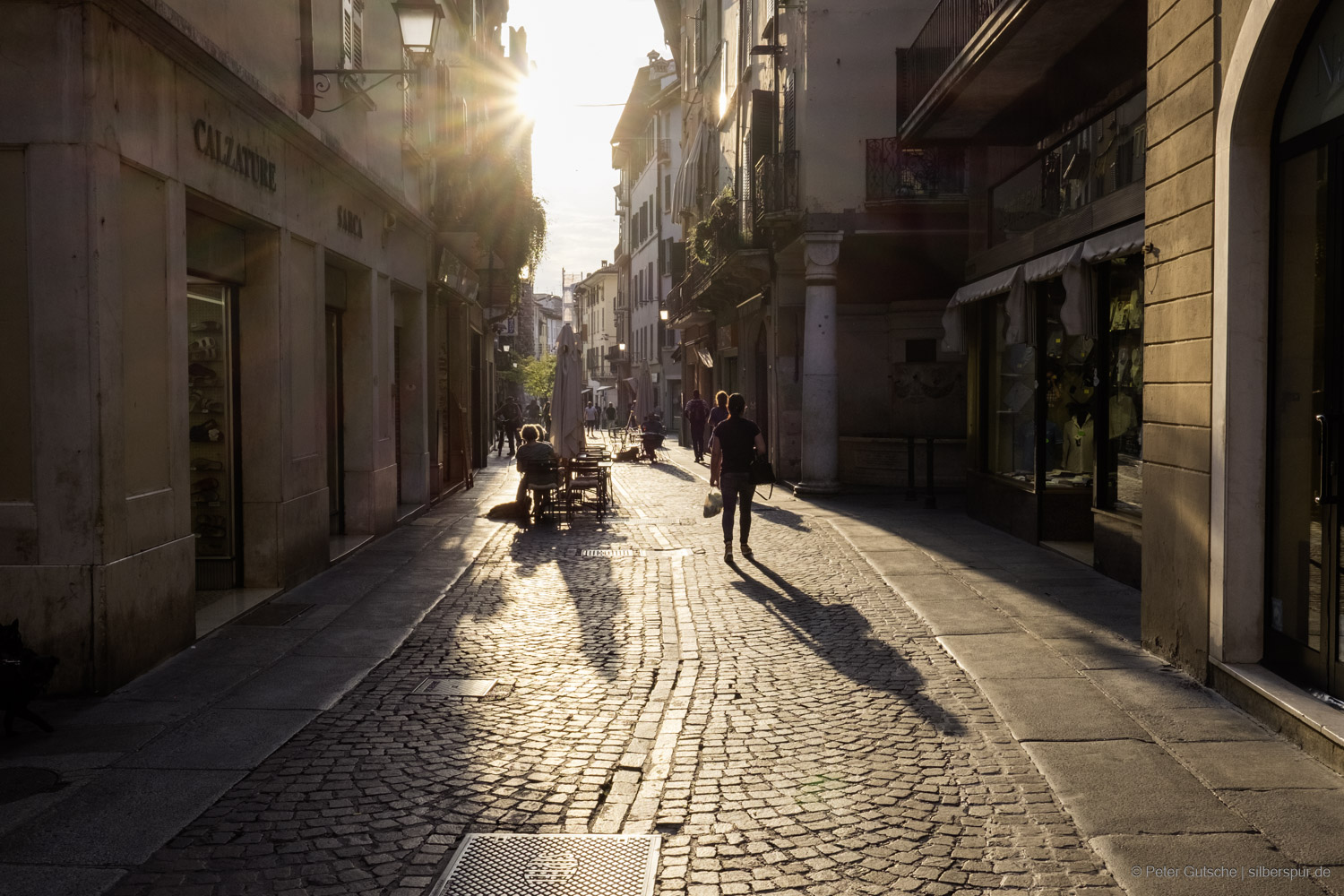 A cobblestone street in a historic old town leading into the low afternoon sun. The sun shines directly into the scene, making the cobblestones gleam. In the backlight, the dark silhouettes of a few pedestrians are visible, walking away from the camera. Further ahead, a table is set up on the street, belonging to a café – two people are sitting there, bathed in the warm light of the sun.