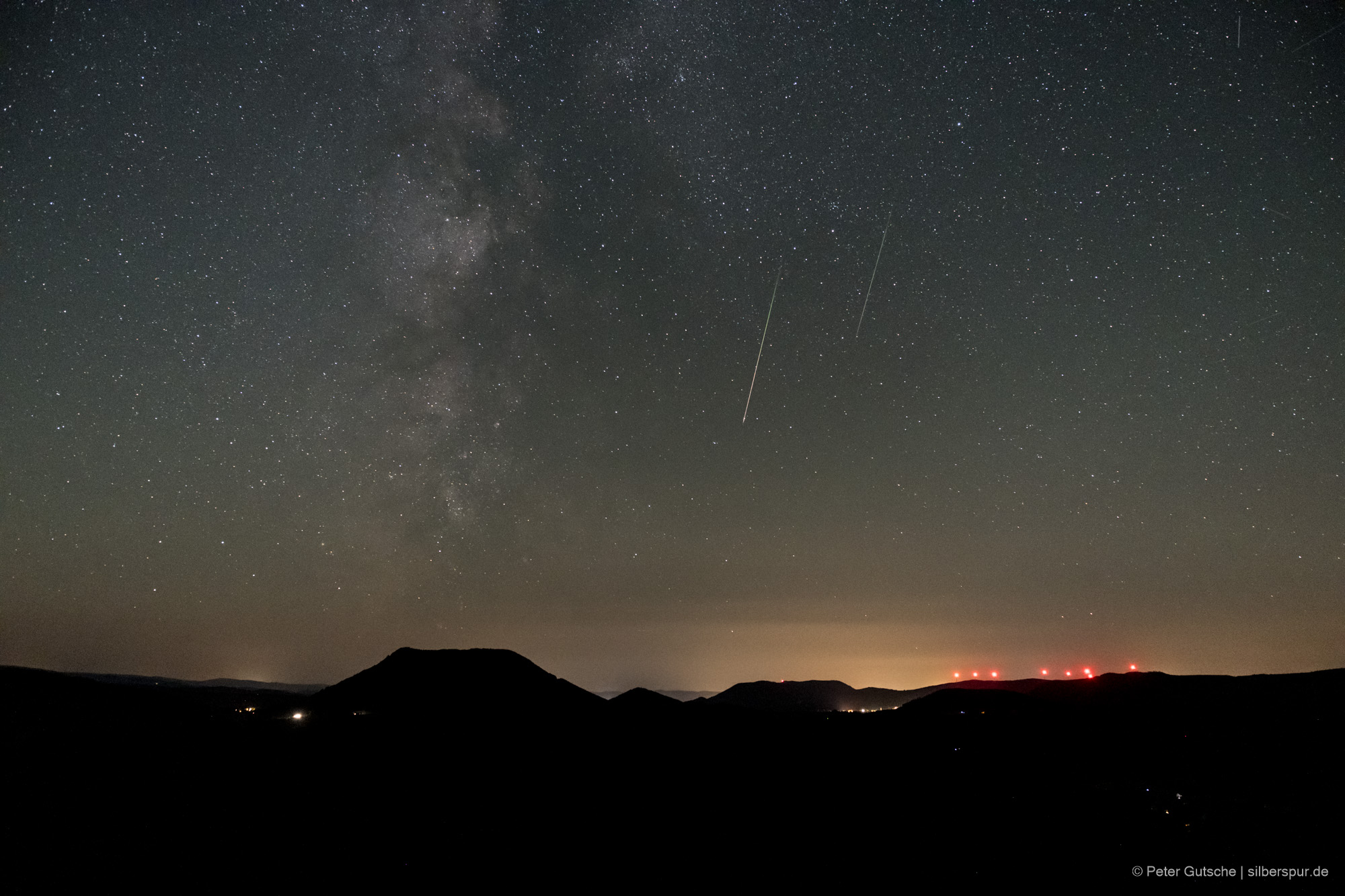 Nachthimmel über einer Berglandschaft. Die dunkle Silhouette eines trapezförmigen Berges (Climont in den Vogesen) hebt sich deutlich von übrigen Landschaftsrelief ab. Darüber der Sternenhimmel und eine einzelne Sternschnuppe, die von oben nach unten im Bild orientiert ist.