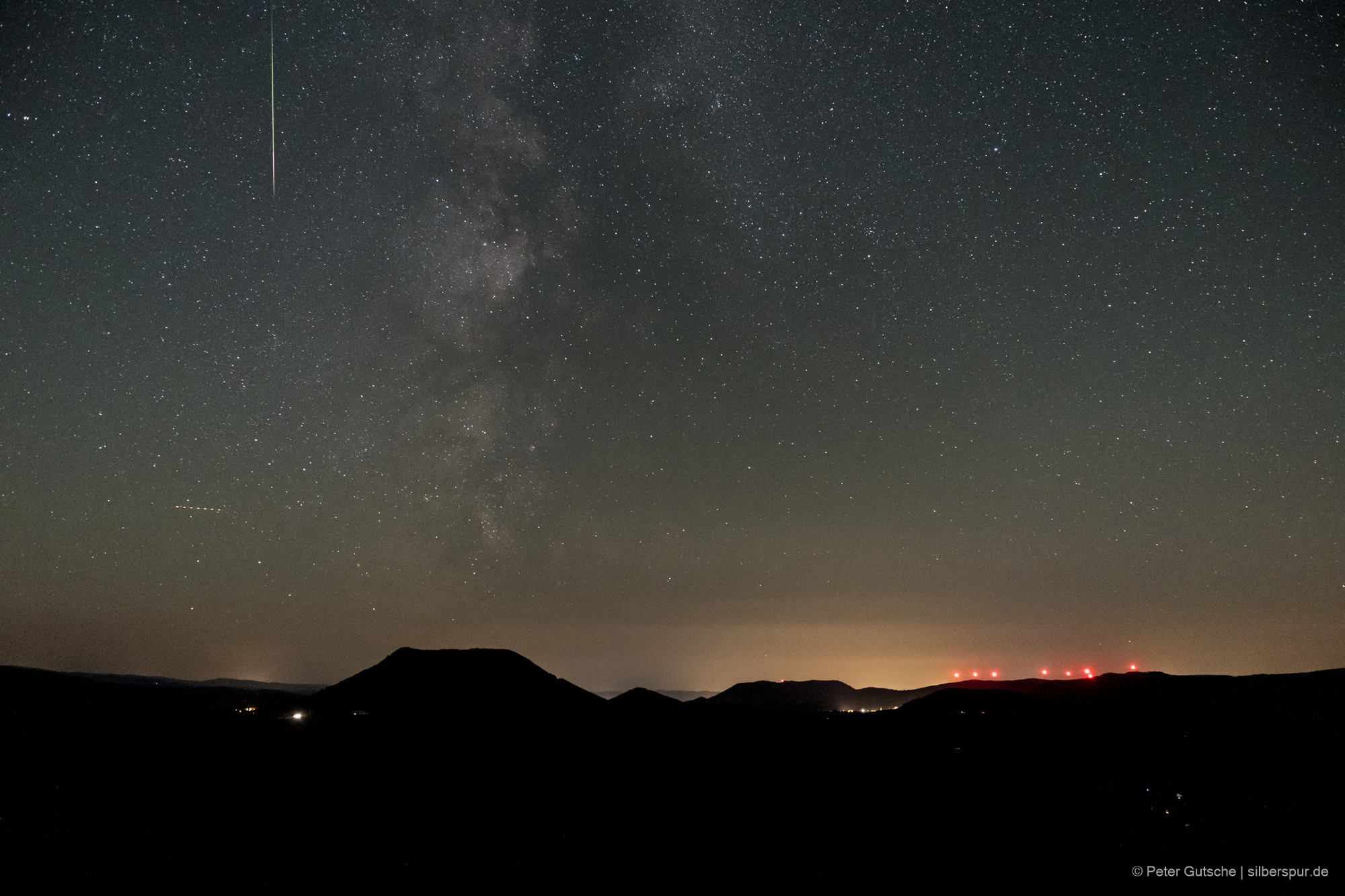 Nachthimmel über einer Berglandschaft. Die dunkle Silhouette eines trapezförmigen Berges (Climont in den Vogesen) hebt sich deutlich von übrigen Landschaftsrelief ab. Darüber der Sternenhimmel und eine einzelne Sternschnuppe, die von oben nach unten im Bild orientiert ist.