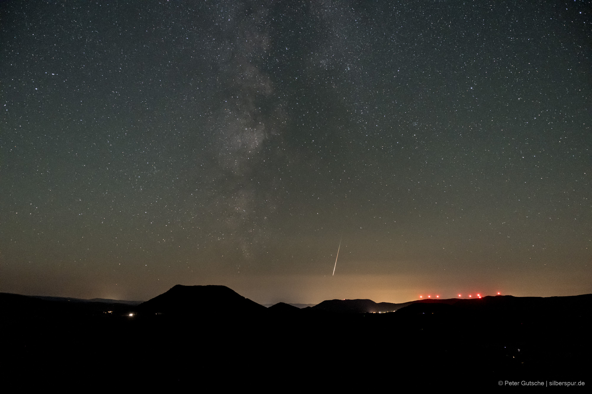 Nachthimmel über einer Berglandschaft. Die dunkle Silhouette eines trapezförmigen Berges (Climont in den Vogesen) hebt sich deutlich von übrigen Landschaftsrelief ab. Darüber der Sternenhimmel und eine einzelne Sternschnuppe, die von oben nach unten im Bild orientiert ist.
