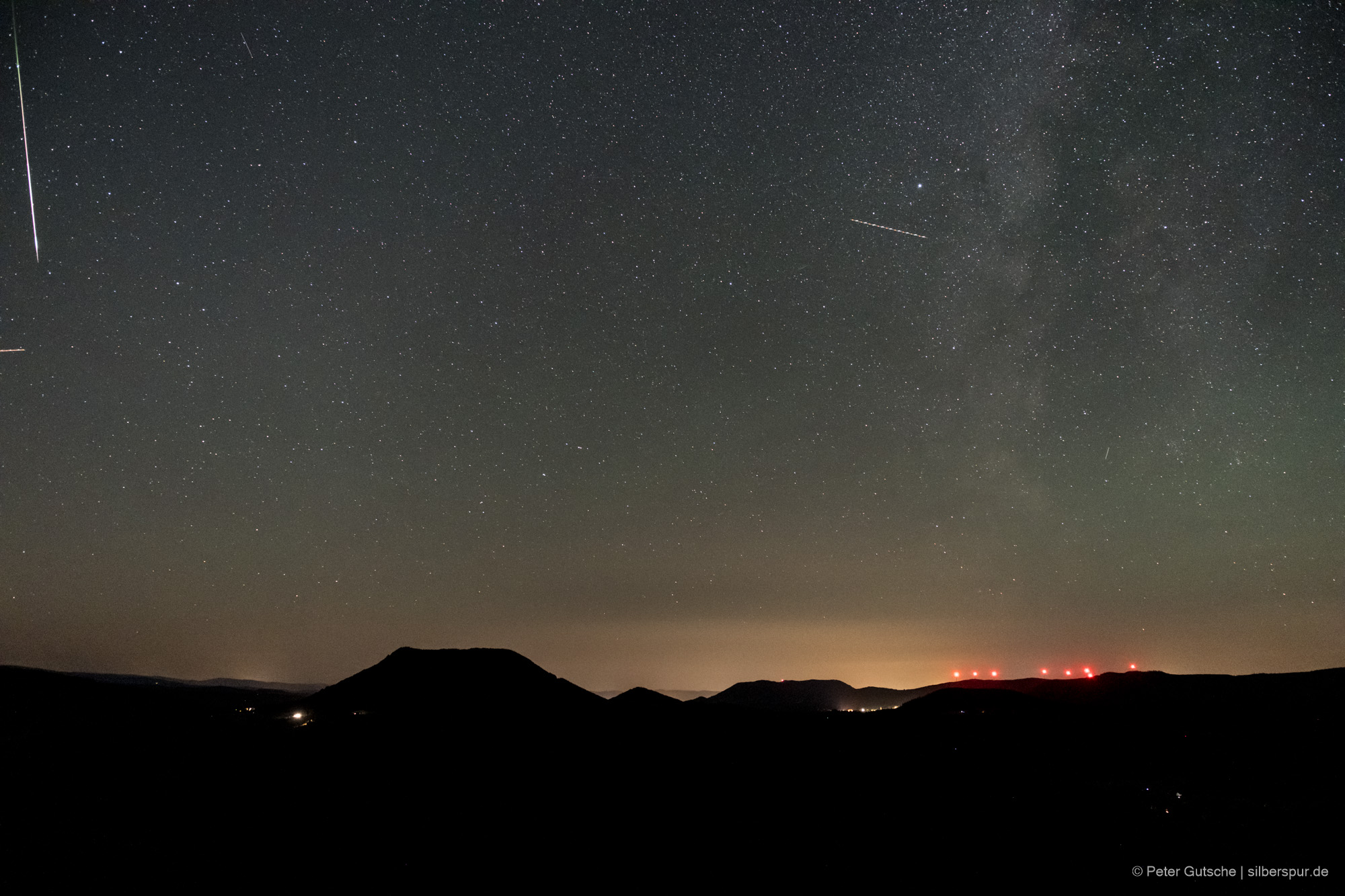 Nachthimmel über einer Berglandschaft. Die dunkle Silhouette eines trapezförmigen Berges (Climont in den Vogesen) hebt sich deutlich von übrigen Landschaftsrelief ab. Darüber der Sternenhimmel und eine einzelne Sternschnuppe, die von oben nach unten im Bild orientiert ist.