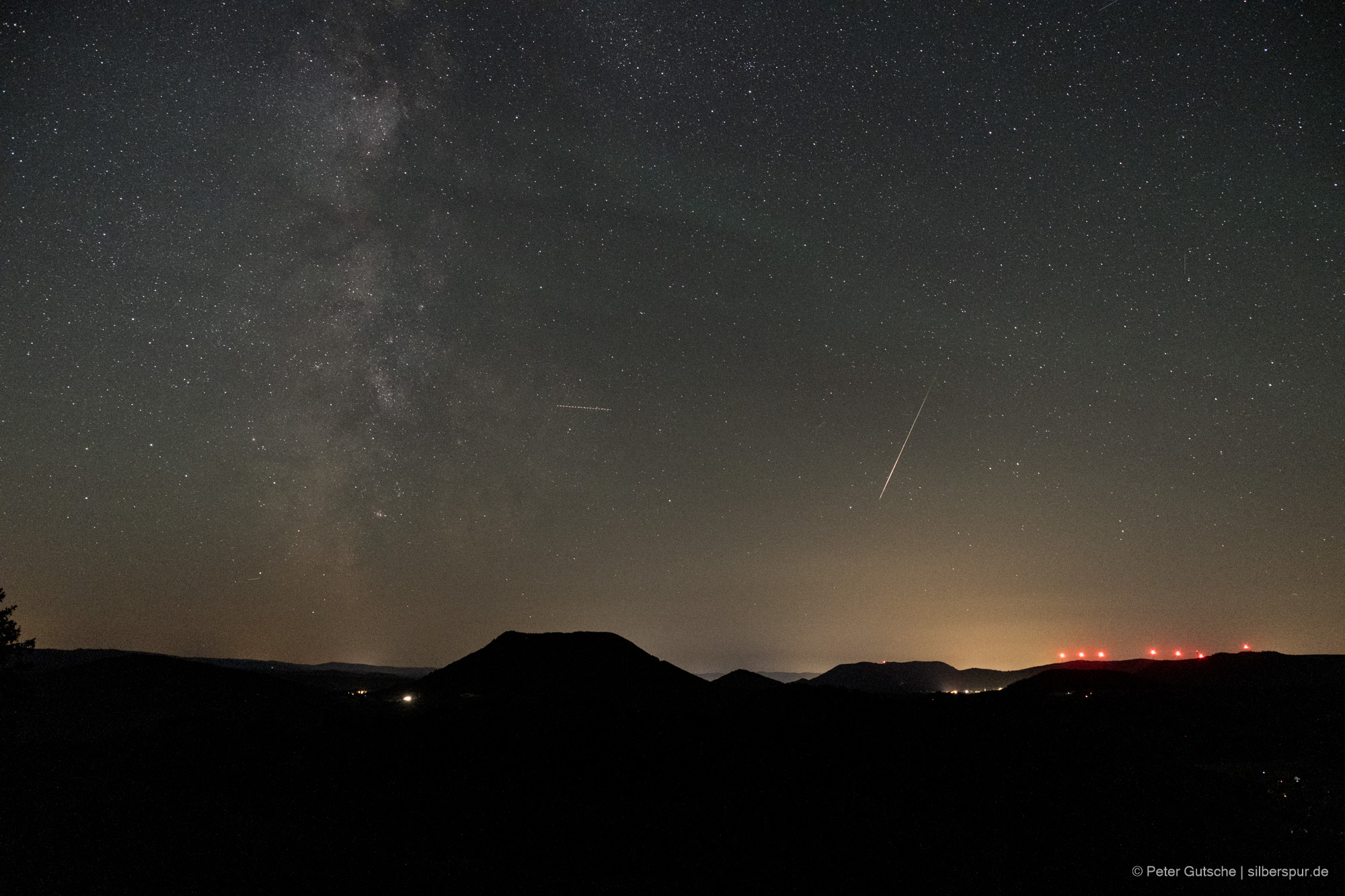 Nachthimmel über einer Berglandschaft. Die dunkle Silhouette eines trapezförmigen Berges (Climont in den Vogesen) hebt sich deutlich von übrigen Landschaftsrelief ab. Darüber der Sternenhimmel und eine einzelne Sternschnuppe, die von oben nach unten im Bild orientiert ist.