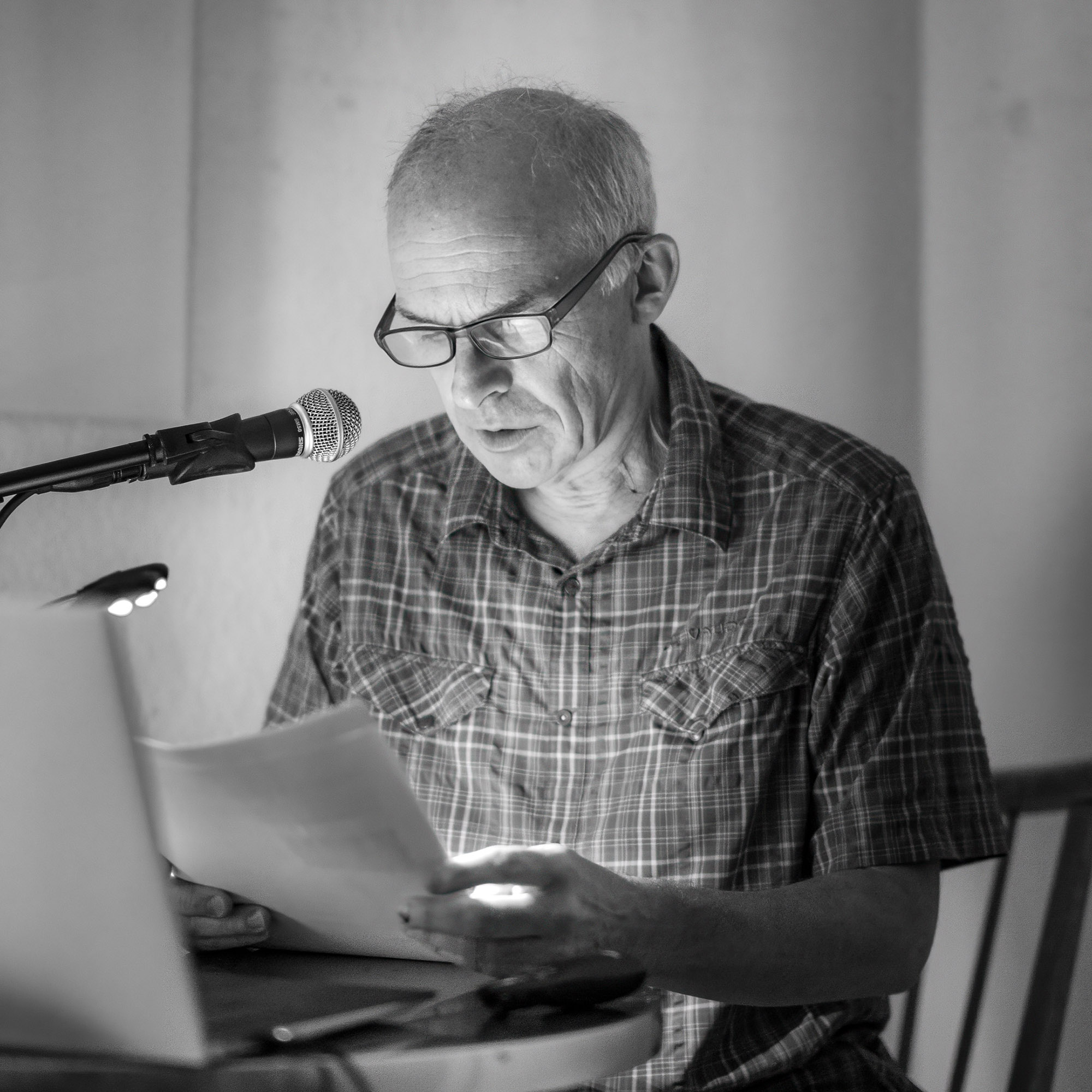 Ein Mann, der, an einem Pult sitzend, einen Text vorliest. Vor ihm auf dem Tisch ein Computer und das Redeskript.