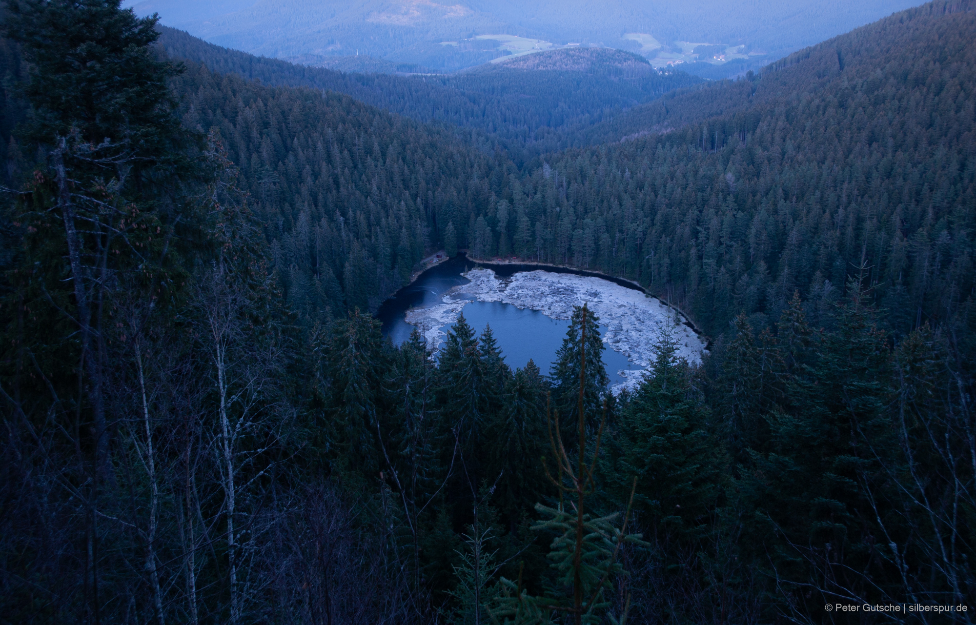Blick auf den fast kreisrunden Huzenbacher See, der zwischen dichten Wäldern in einer Mulde liegt. Der See ist zu Teilen von Moor bedeckt, das hier von Raureif bedeckt ist. Das Foto wurde am späten Nachmittag gemacht, weshalb der See im Schatten liegt.