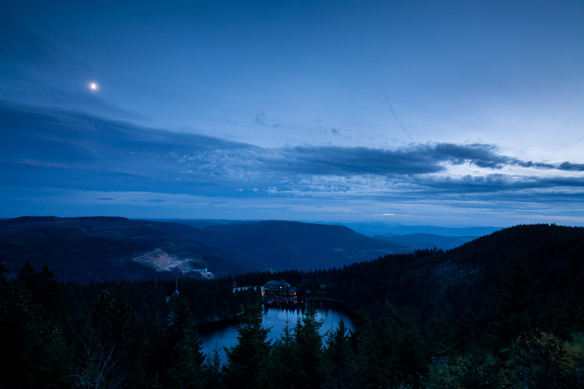 An einem Abend, ein See von einem Berg aus fotografiert, an dessen Ufer ein Hotel steht. In der Dämmerung ist alles in blaue Farbe getaucht, und aus den Hotelfenstern leuchtet das Licht. Der See liegt hoch oberhalb, so dass man hinter dem See weiter in die Tiefe und im Hintergrund auf den südlichen Schwarzwald schaut. Am Himmel ziehen Wolken auf, dennoch ist die Mondsichel sichtbar.