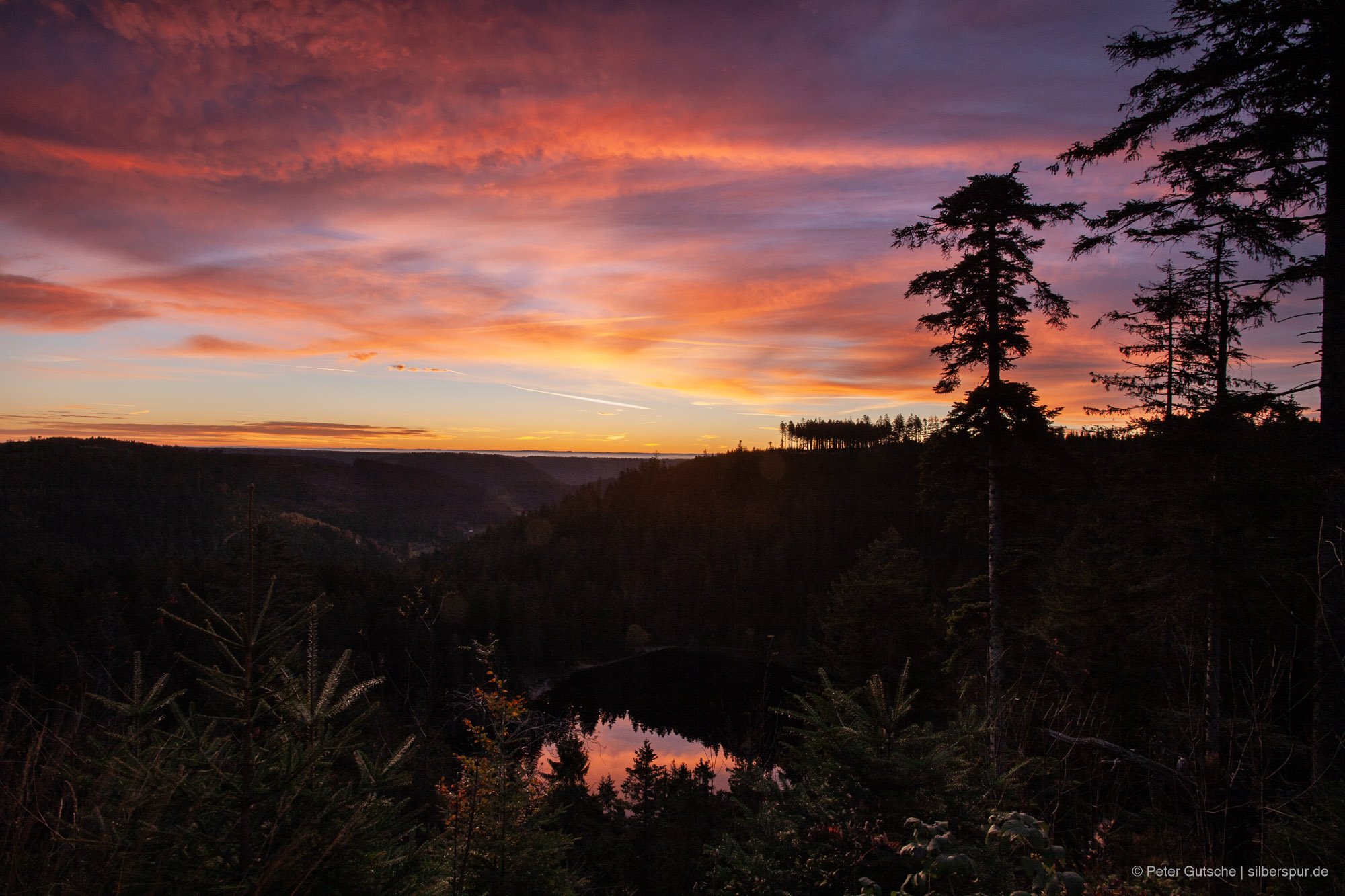Ein in einer Waldsenke von dichtem Nadelwald umgebener See in der Morgendämmerung. Die umliegenden Berge sind schwarze Schatten, während der See das glutrote Licht des Himmels widerspiegelt.