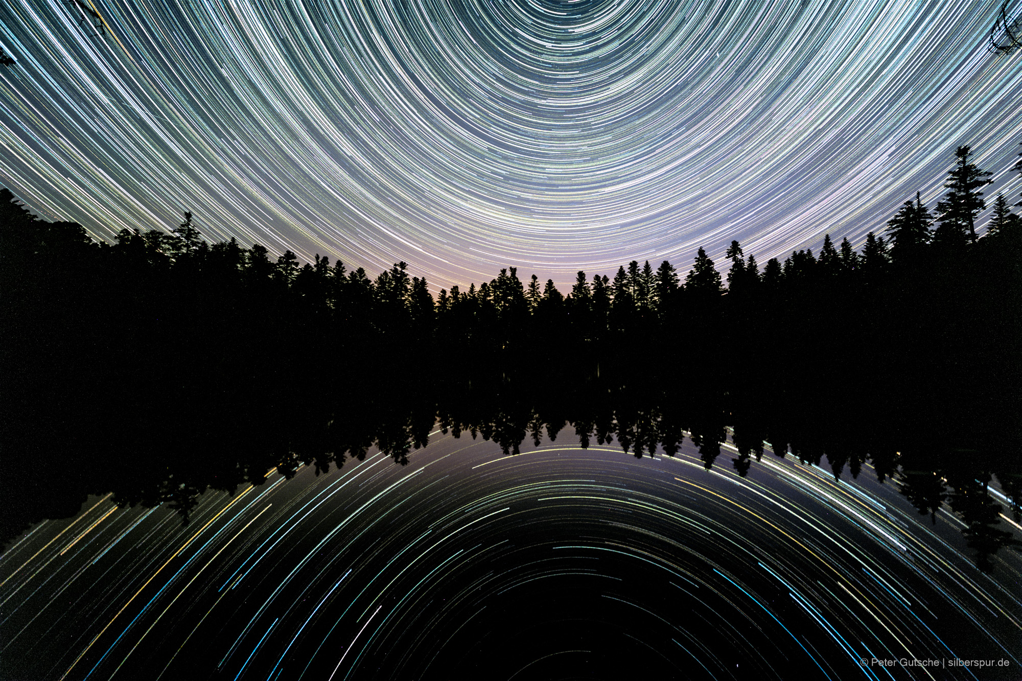 Star trails are reflected in a lake at night. Since the photo was taken directly facing north and the horizon, with the row of trees on the opposite shore, was placed in the center, the star trails form two arcs that are symmetrically mirrored.