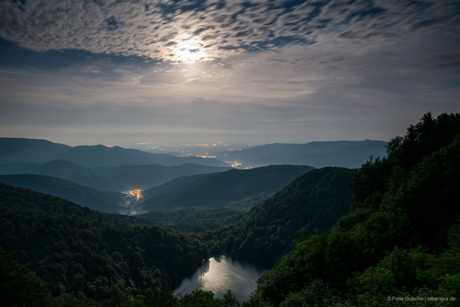 A lake seen from above at night with the full moon in the sky (slightly dimmed by high altitude clouds) and the moon light reflected on the lake surface. In the backgriund, a mountain landscape with valleys. In the valleys, the lights of smaller villages illuminate the night.