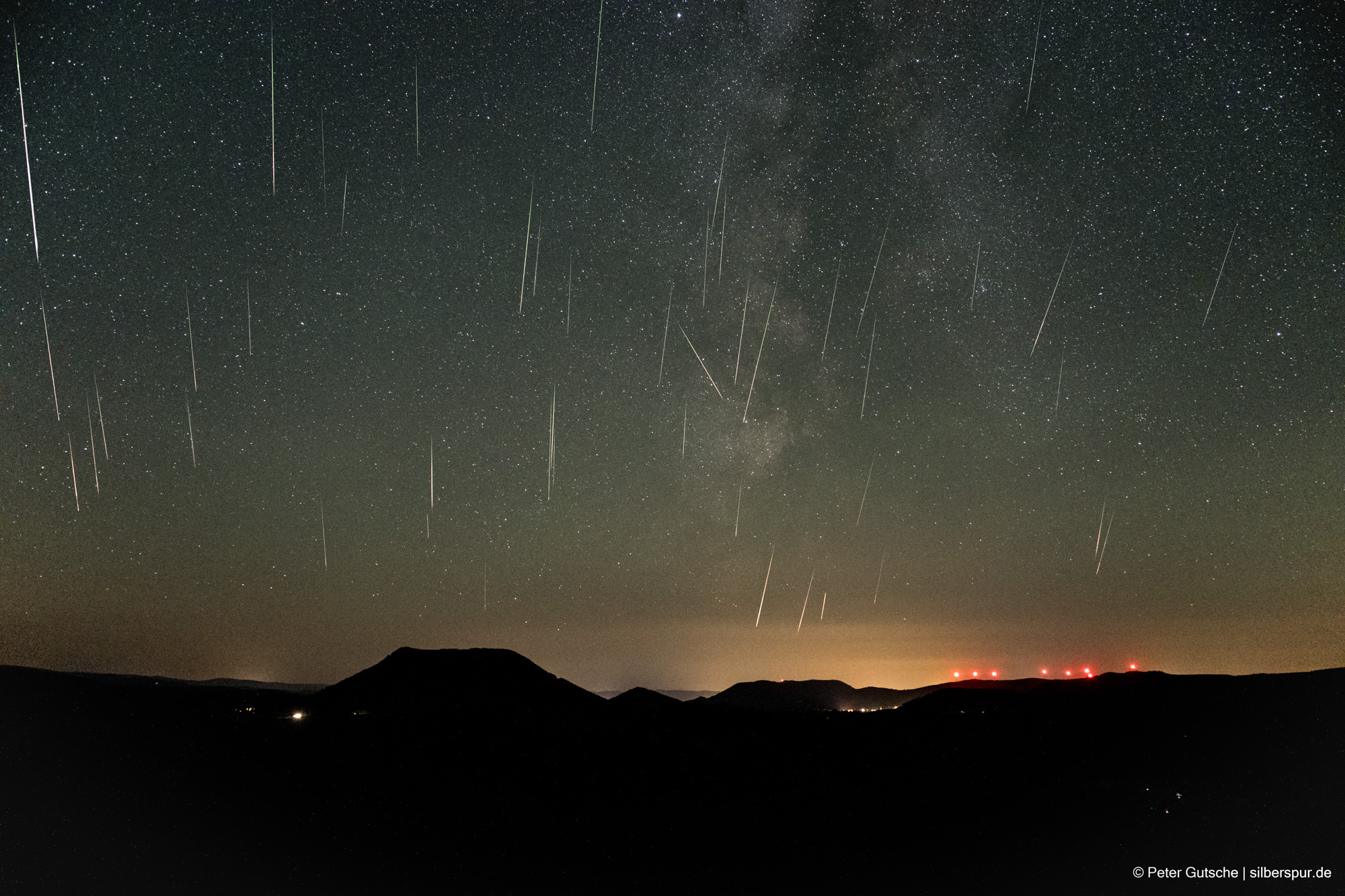 Nachthimmel über einer Berglandschaft. Es zeigt den Nachthimmel über einer Berglandschaft mit Sternen, der Milchstraße und zahlreichen Meteoren, die scheinbar von oben nach unten fallen. Die dunkle Silhouette eines trapezförmigen Berges (Climont in den Vogesen) hebt sich deutlich von übrigen Landschaftsrelief ab.